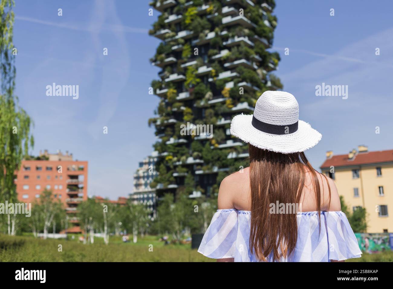 Girl tourist with a sun hat looking at a skyscraper with plant and tree-covered balconies and terraces. Architecture, travel, and lifestyle concepts. Stock Photo