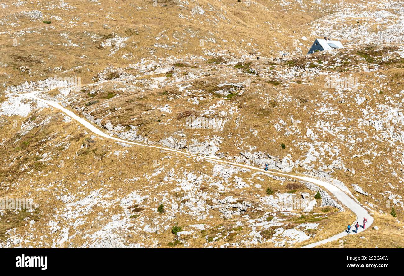 Hikers ascending a winding mountain path towards a mountain hut nestled amidst a vast, rocky landscape. Mount mangart, slovenia Stock Photo