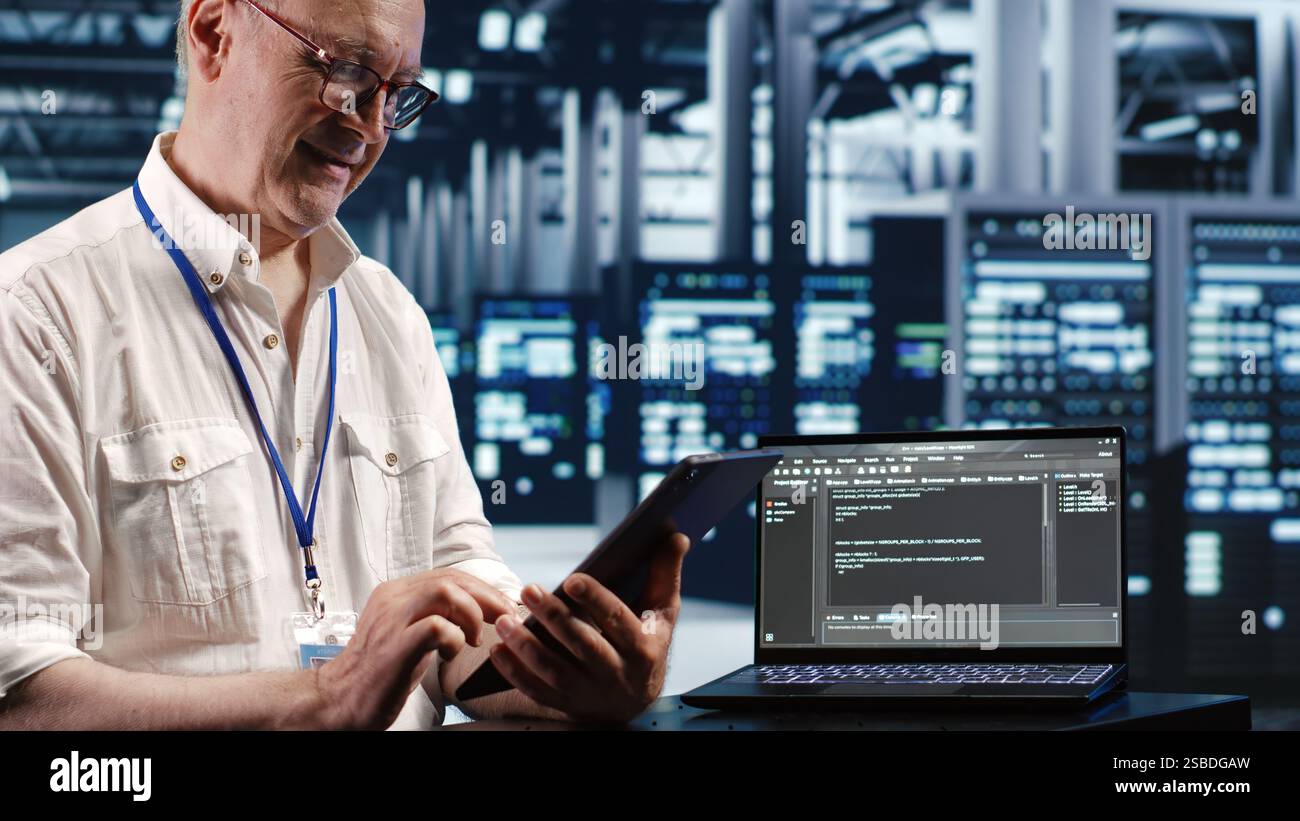 Elderly worker in data center, solving tasks on tablet while running scripts on laptop terminal. Certified engineer amidst servers, accessing databases and manipulating lines of code Stock Photo