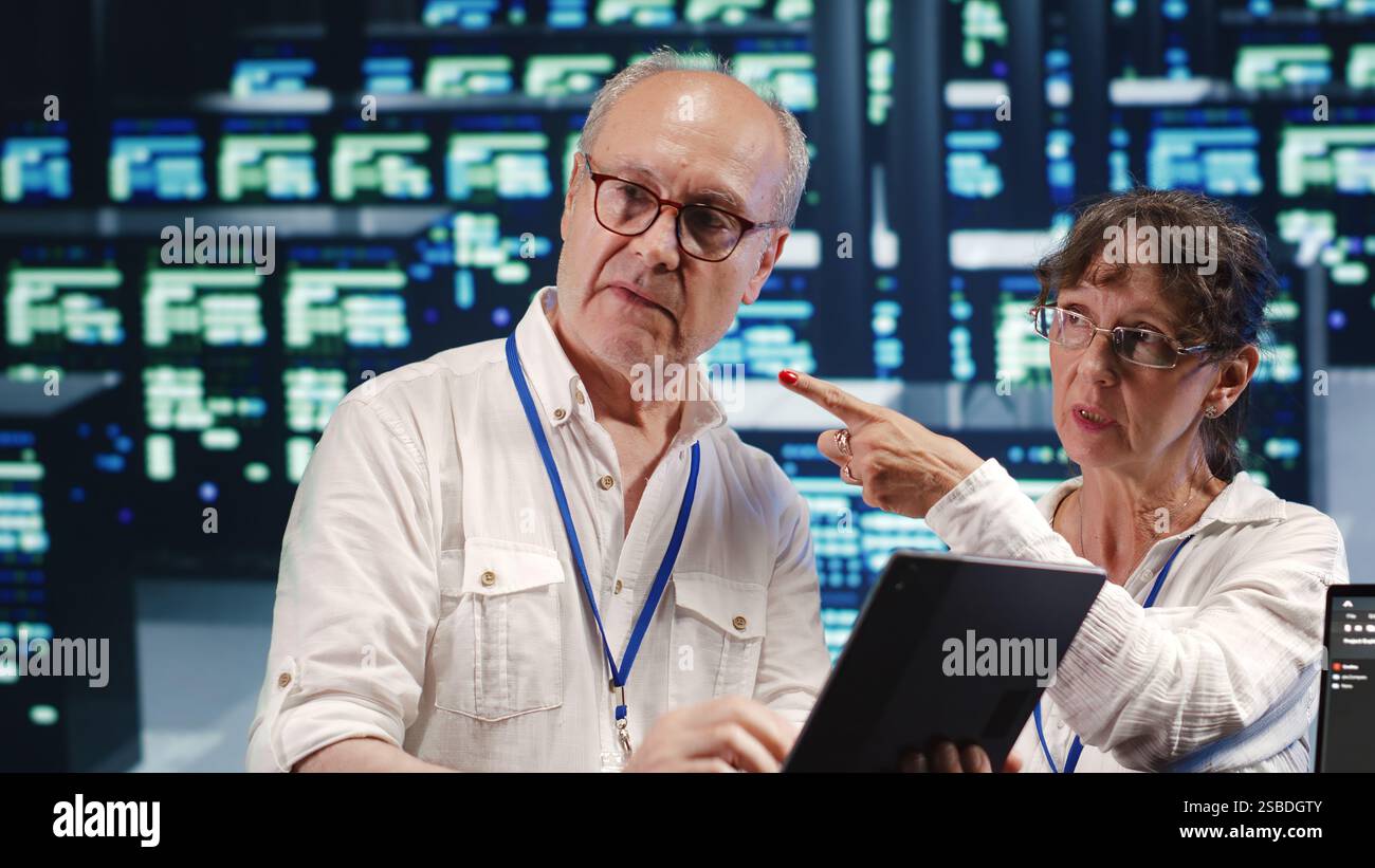 Software developers in high tech establishment, completing tasks on tablet while running scripts on laptop terminal. Proactive programmers amidst server rows, accessing databases and manipulating code Stock Photo