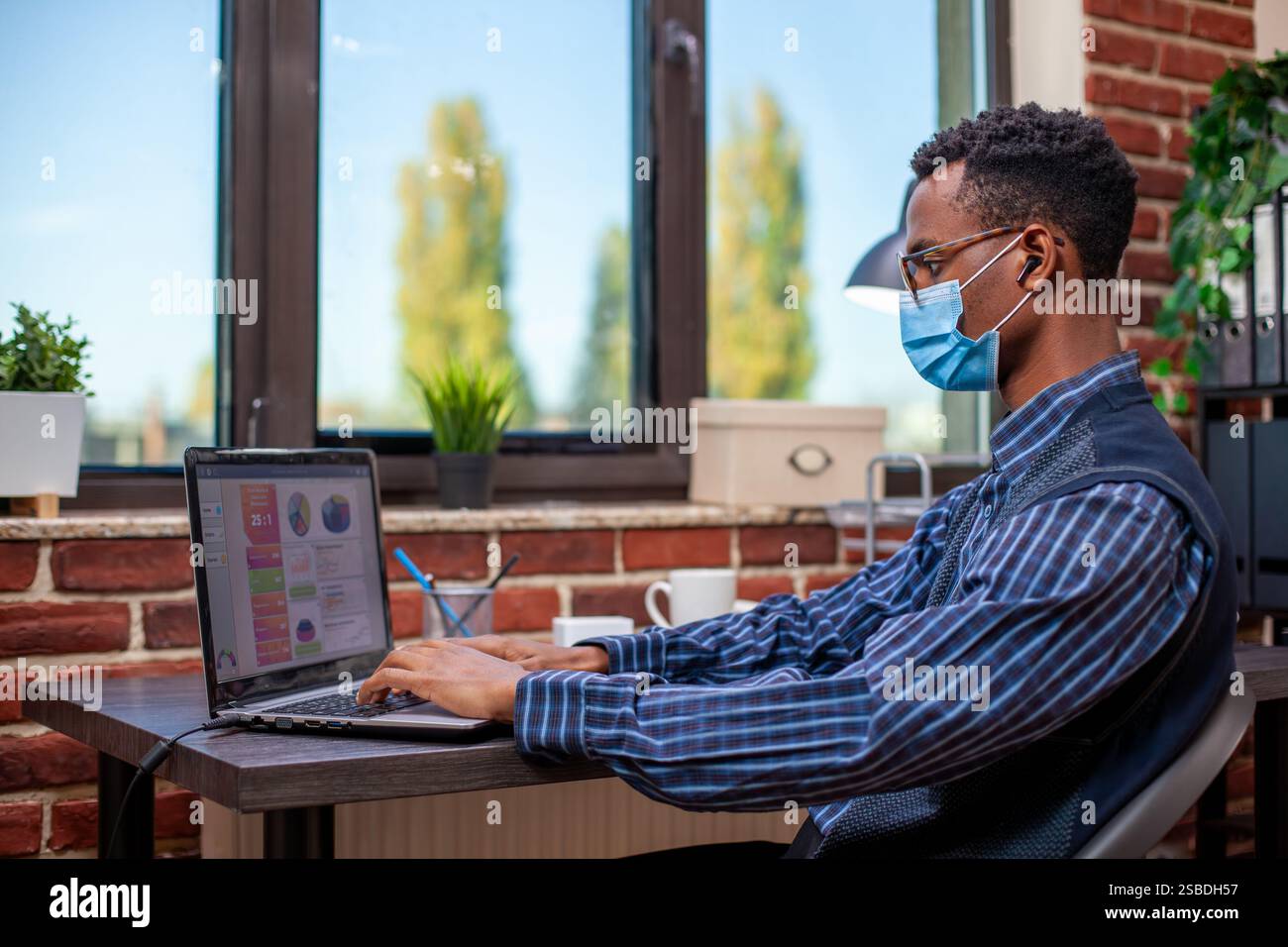 Focused manager works in startup office typing on his laptop and reviewing online documents, to improve company growth. Male project analyst with face mask and wireless earbuds, working at his desk. Stock Photo