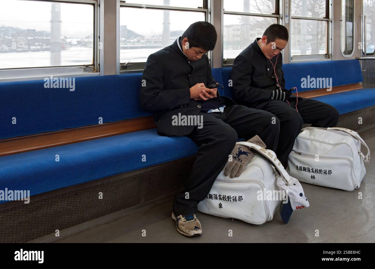 Two high school boys wearing matching traditional "gakuran" (black uniforms) looking at cell phones and carrying sports bags riding a train in Japan. Stock Photo