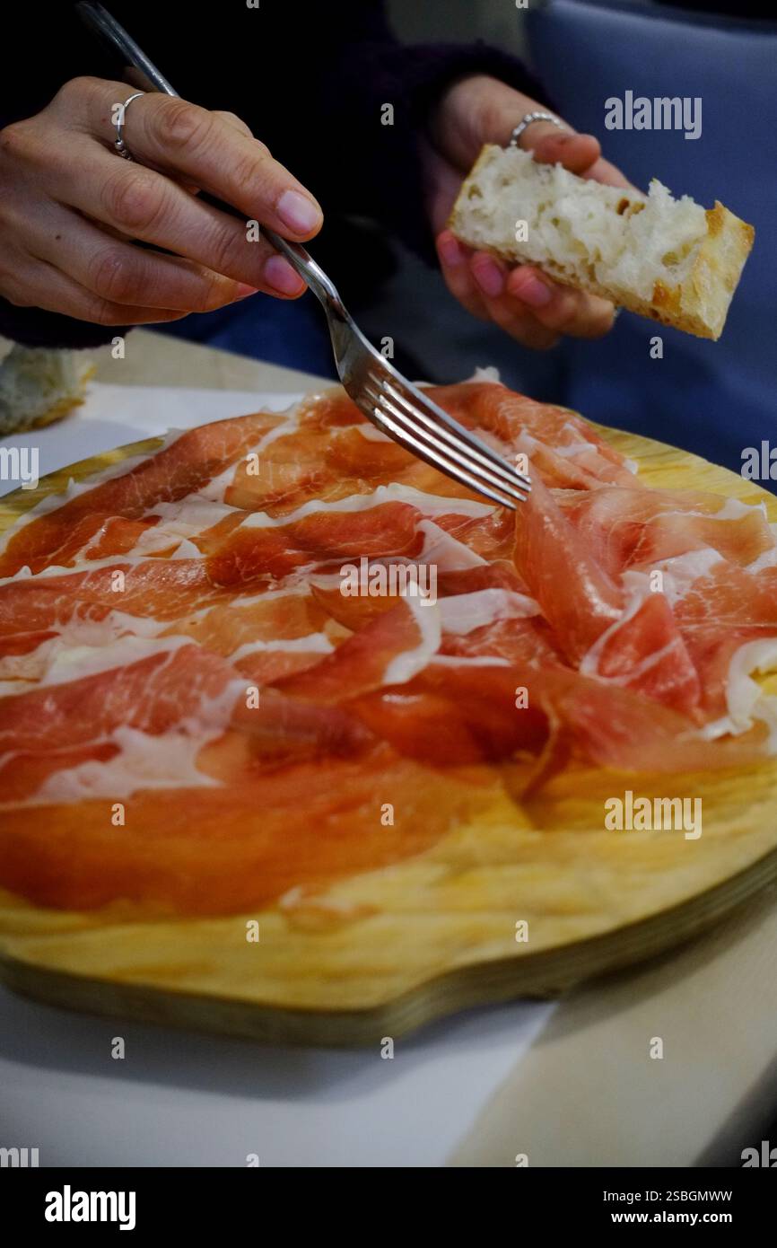 Thinly sliced cured meat arranged on a wooden board, alongside a piece of crusty bread. A person uses a fork to prepare a bite. Stock Photo