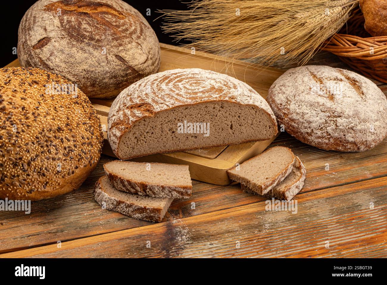 Different kinds of bread are presented on a wooden table with wheat ears, creating a rustic bakery atmosphere Stock Photo