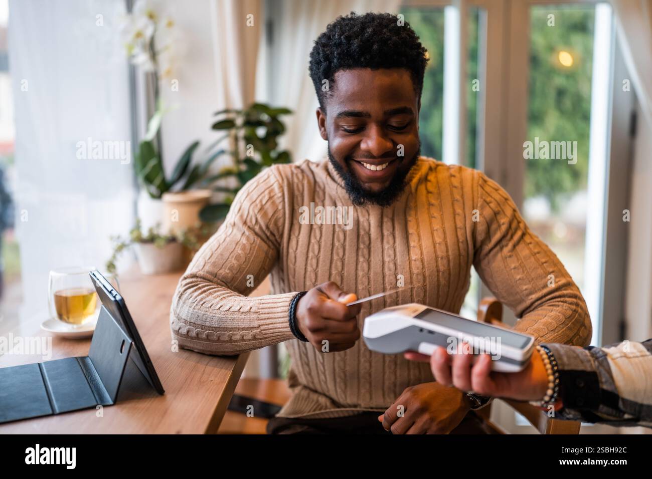 Young man in casual clothing makes a contactless payment with payment card while sitting in the cozy city cafe. Stock Photo