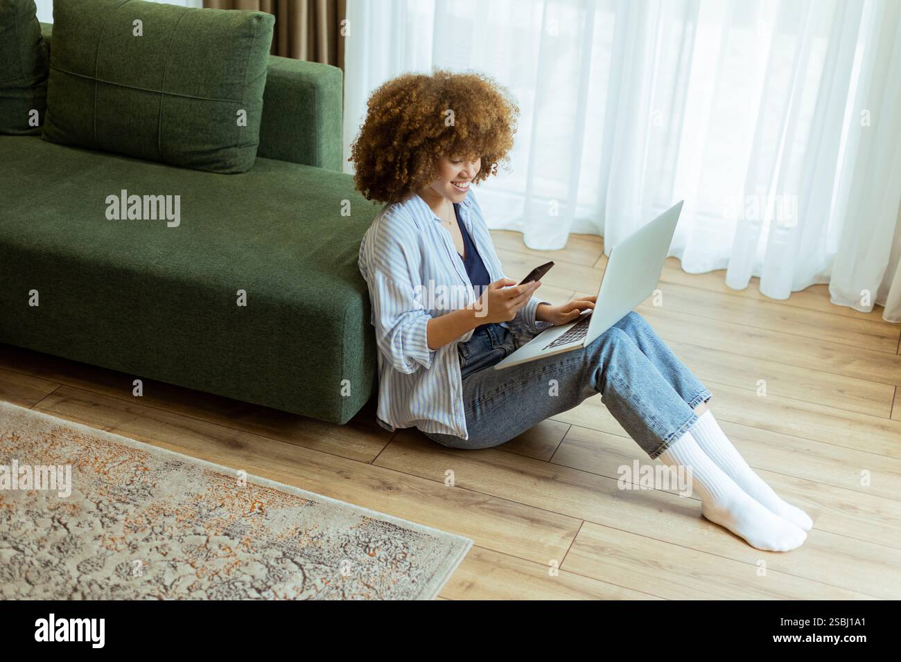 Seated comfortably on the floor, a young woman engages with her laptop while checking her smartphone. The cozy, sunlit living room creates a serene ba Stock Photo