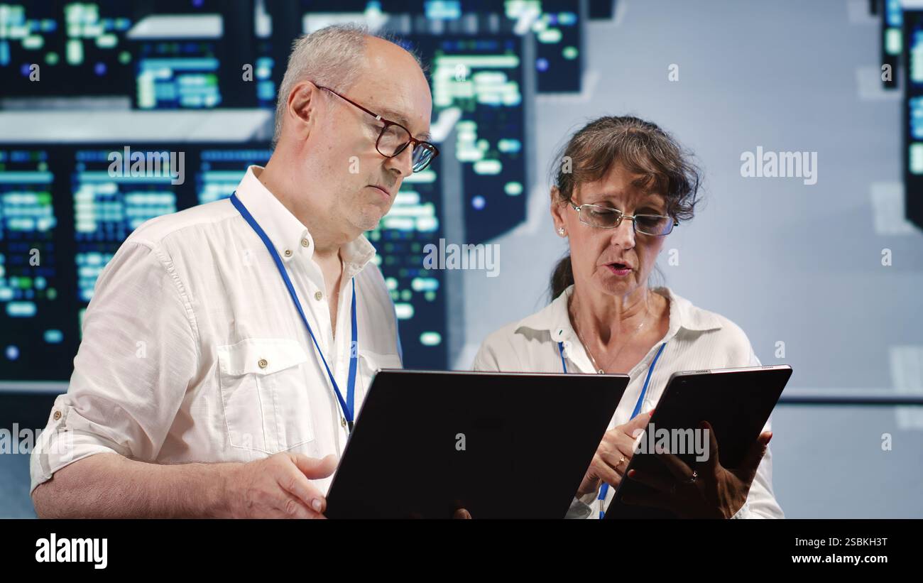 Data storage company executives looking around data center, using laptop and tablet to crosscheck contingency plan and assess high tech workspace units in need of fixing, preventing liabilities Stock Photo
