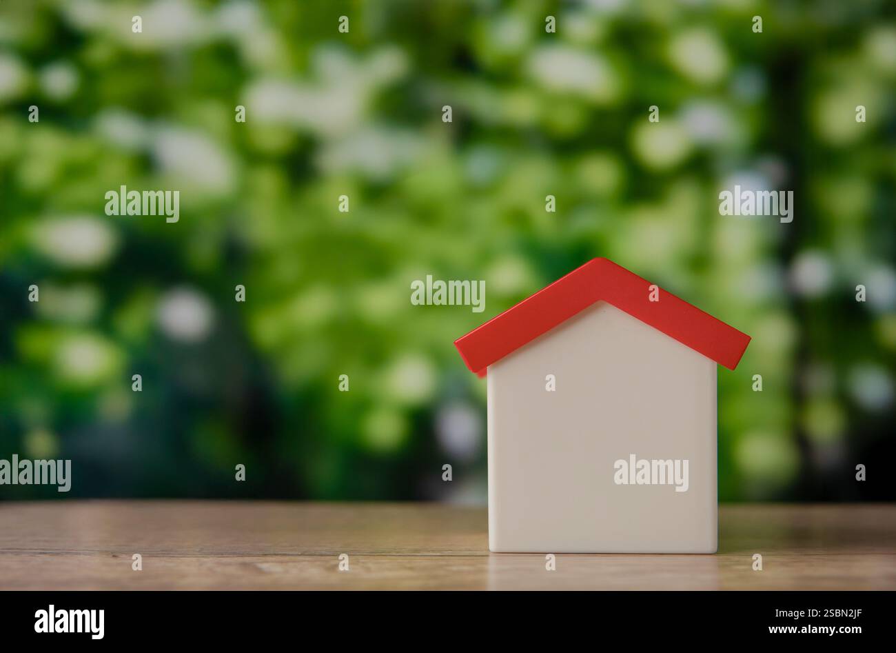 A house model on a wooden table against a blurred background of green foliage, symbolizing a green home or eco-friendly living. Copy space for text. Stock Photo