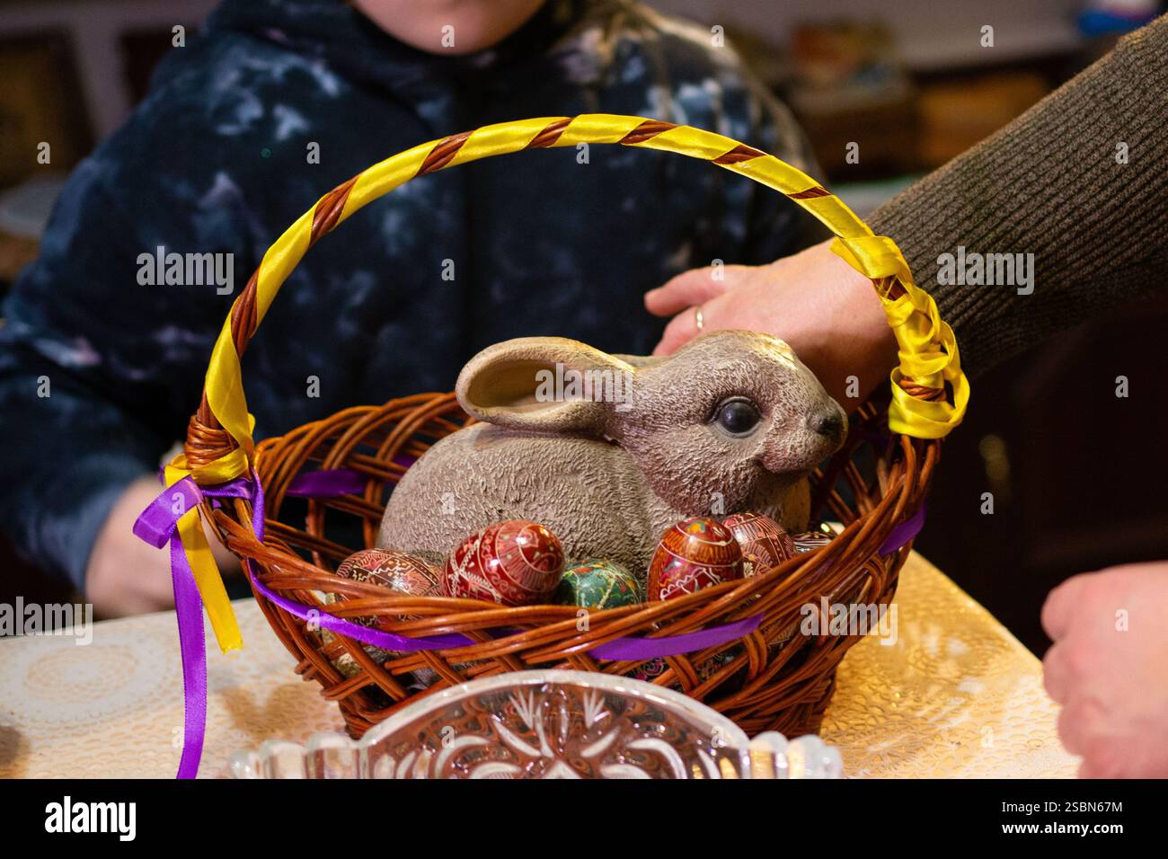 A brown ceramic rabbit rests in a woven basket alongside several intricately decorated eggs. The basket is adorned with yellow and purple ribbons. A p Stock Photo