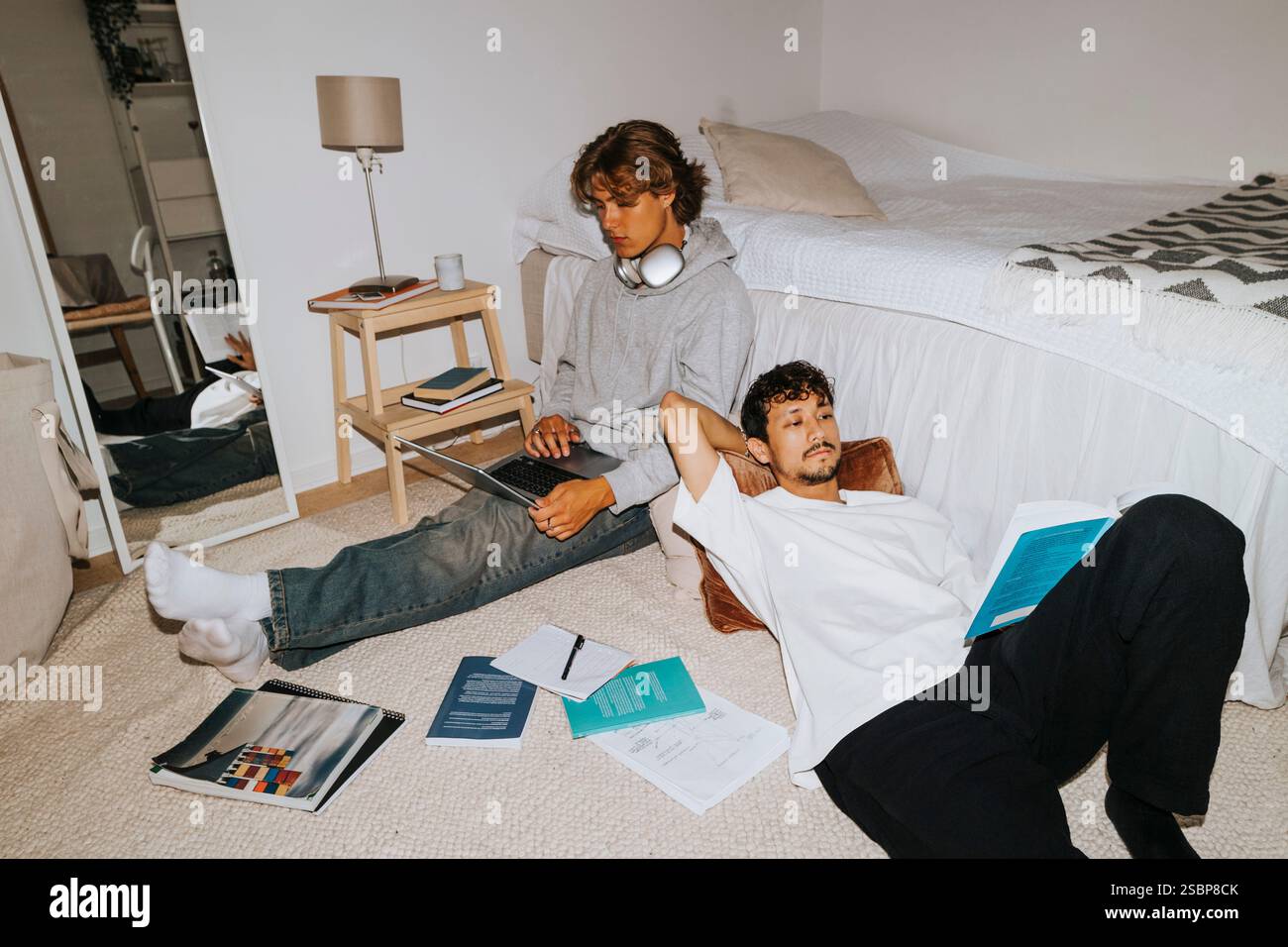 Young man reading book while lying down near male friend using laptop in college dorm bedroom Stock Photo