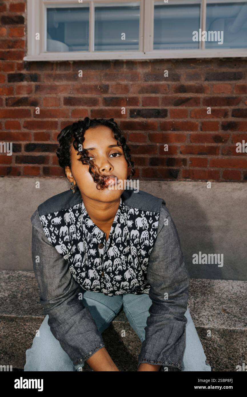 Portrait of young woman posing while sitting on bench against brick wall Stock Photo