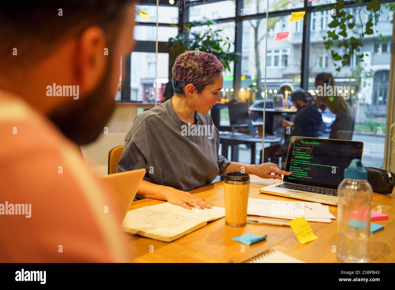 Non-binary IT professional discussing coding on laptop to colleagues during meeting in office Stock Photo
