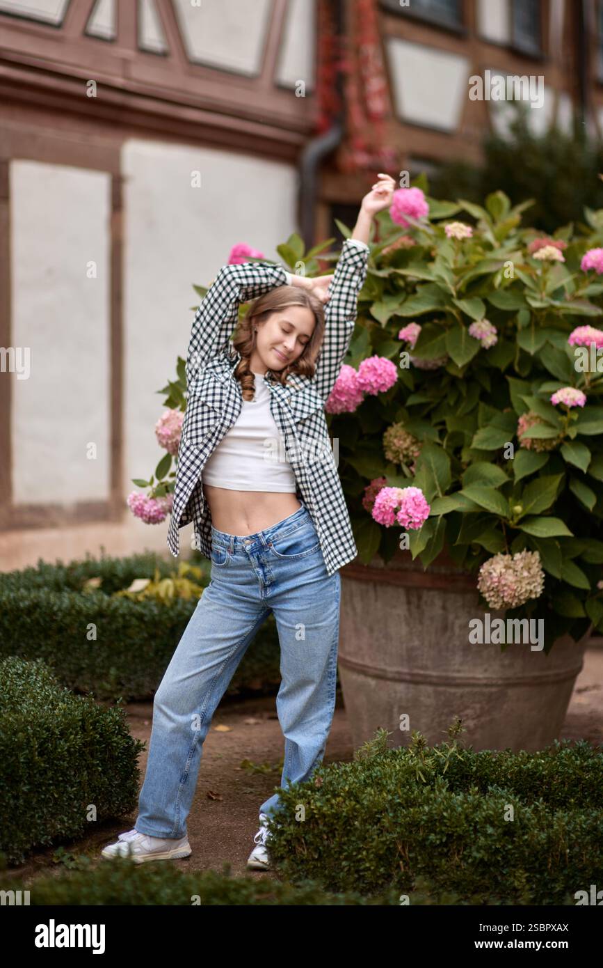 Young woman in casual autumn outfit posing among blooming hydrangeas in front of traditional half-timbered house in Germany – cozy and stylish Stock Photo