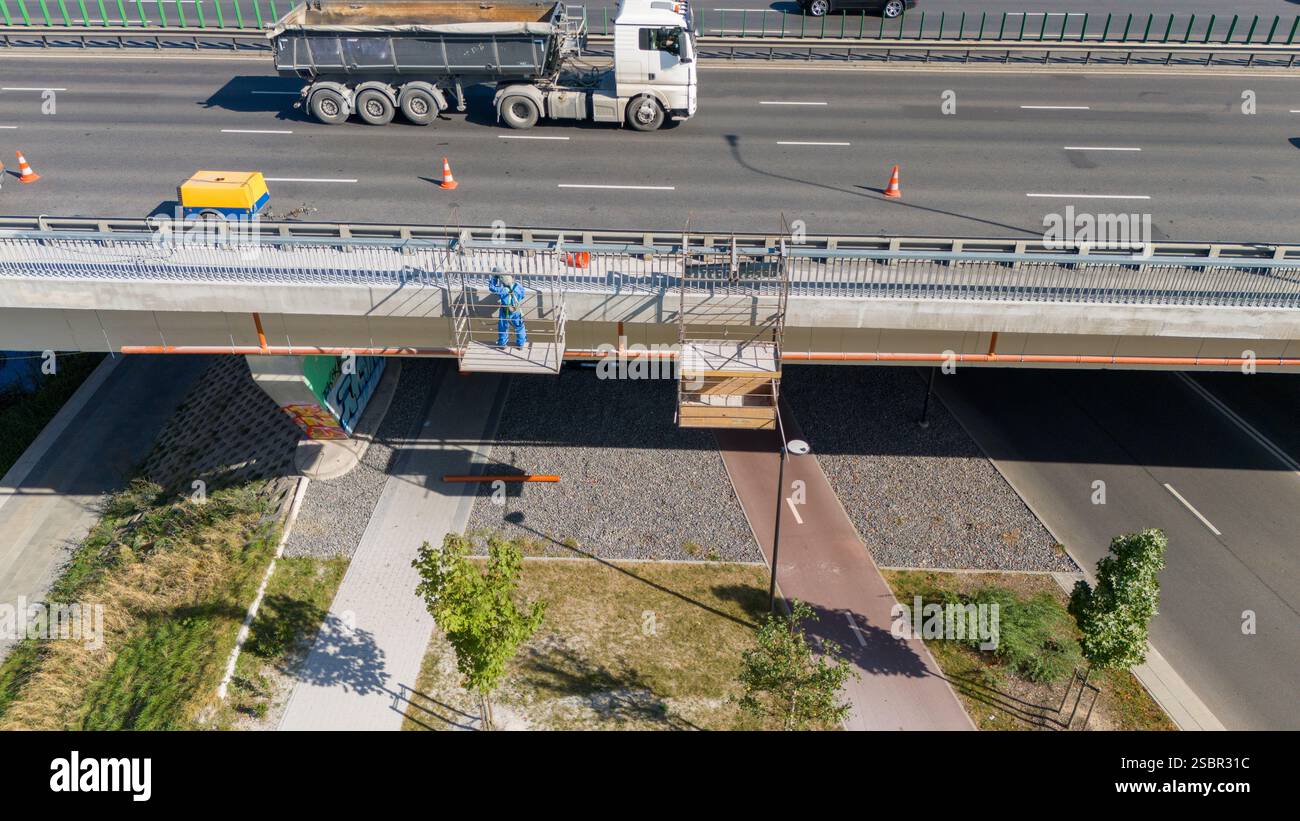 Aerial view of a worker on a suspended platform under a highway bridge, with a truck passing above. The scene includes traffic cones, a bicycle path, Stock Photo