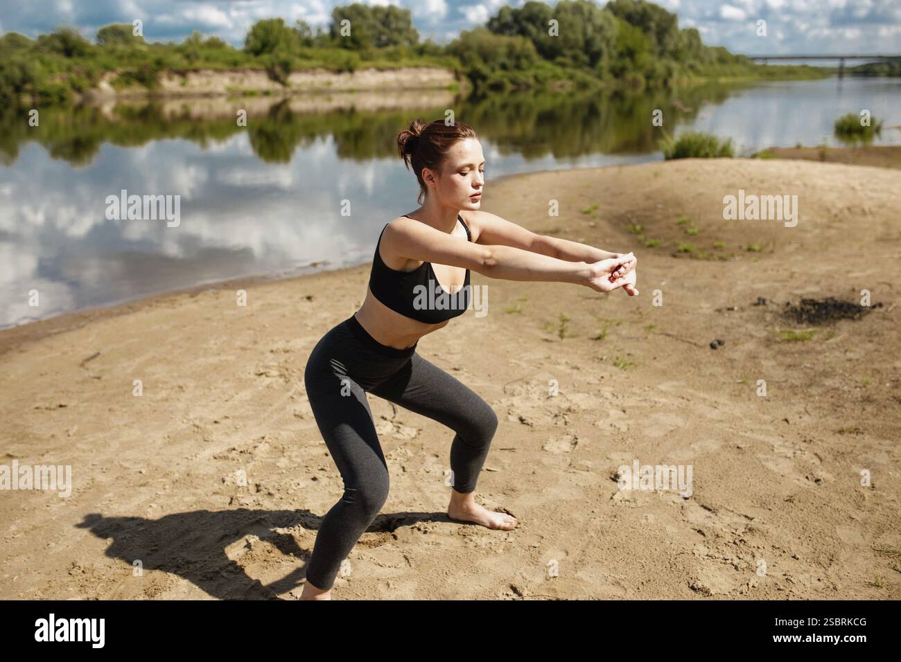 Woman performs a squat exercise on a sandy riverbank, holding a ...