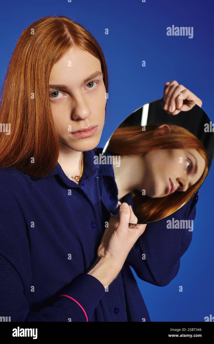 A young man with long red hair gazes thoughtfully into a mirror ...