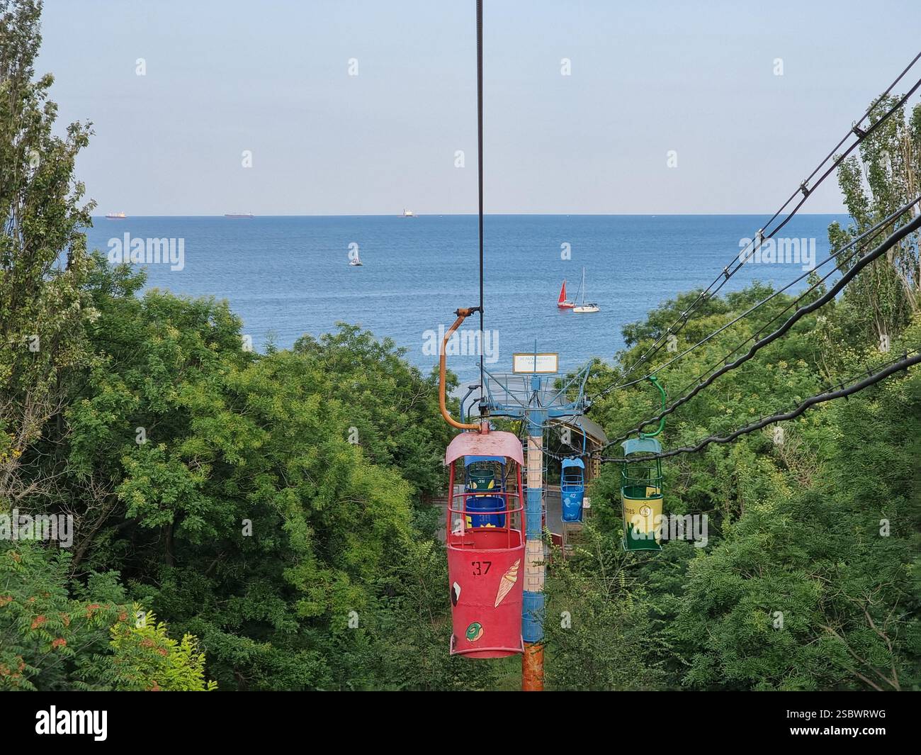 Colourful gondolas of the vintage Soviet cable car in Odesa / Odessa, Ukraine to Otrada beach over forest trees w/ view on Black Sea w/ sailing boats - Smartphone Captured Stock Image