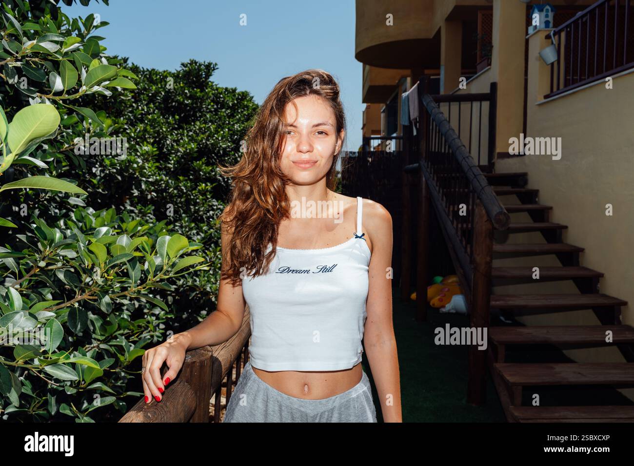 A cheerful young woman poses confidently outdoors by a wooden staircase, surrounded by lush greenery under the bright sun Stock Photo