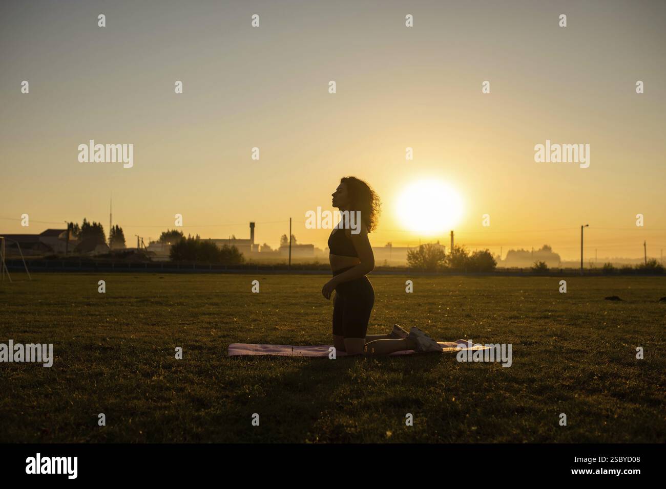 Person kneels on a yoga mat in a meditative pose during sunrise, creating a serene silhouette Stock Photo