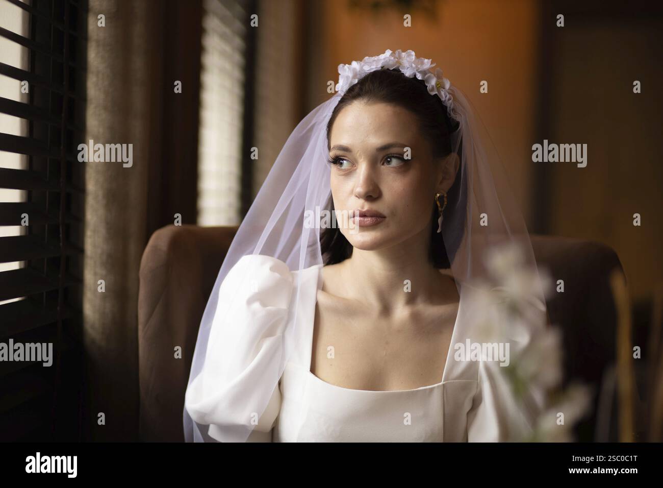 Bride looks introspective sitting by window, soft light filtering through blinds Stock Photo