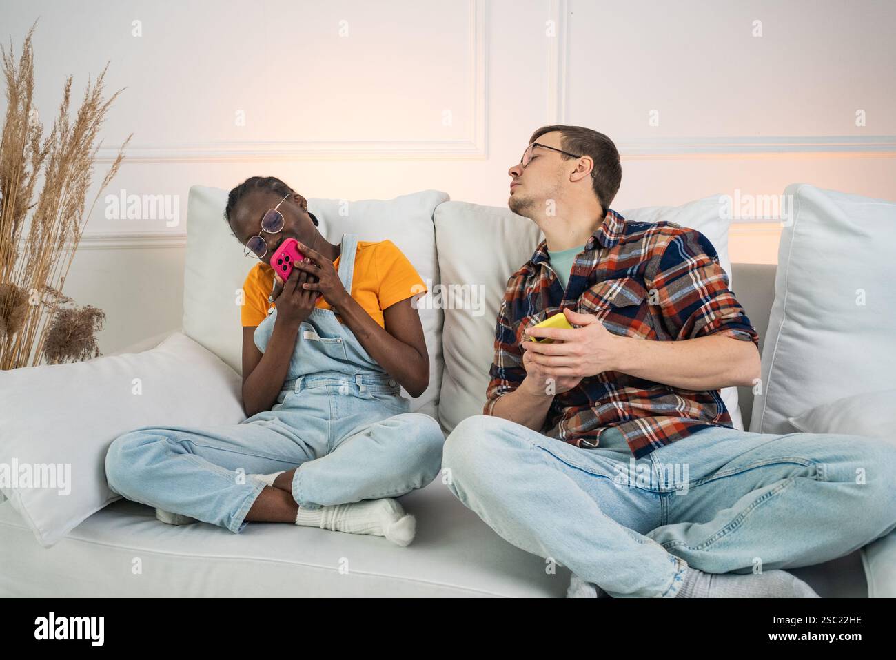 Multiethnic couple sitting on a white sofa, ignoring each other while using their smartphones, highlighting the impact of technology on modern relationships Stock Photo