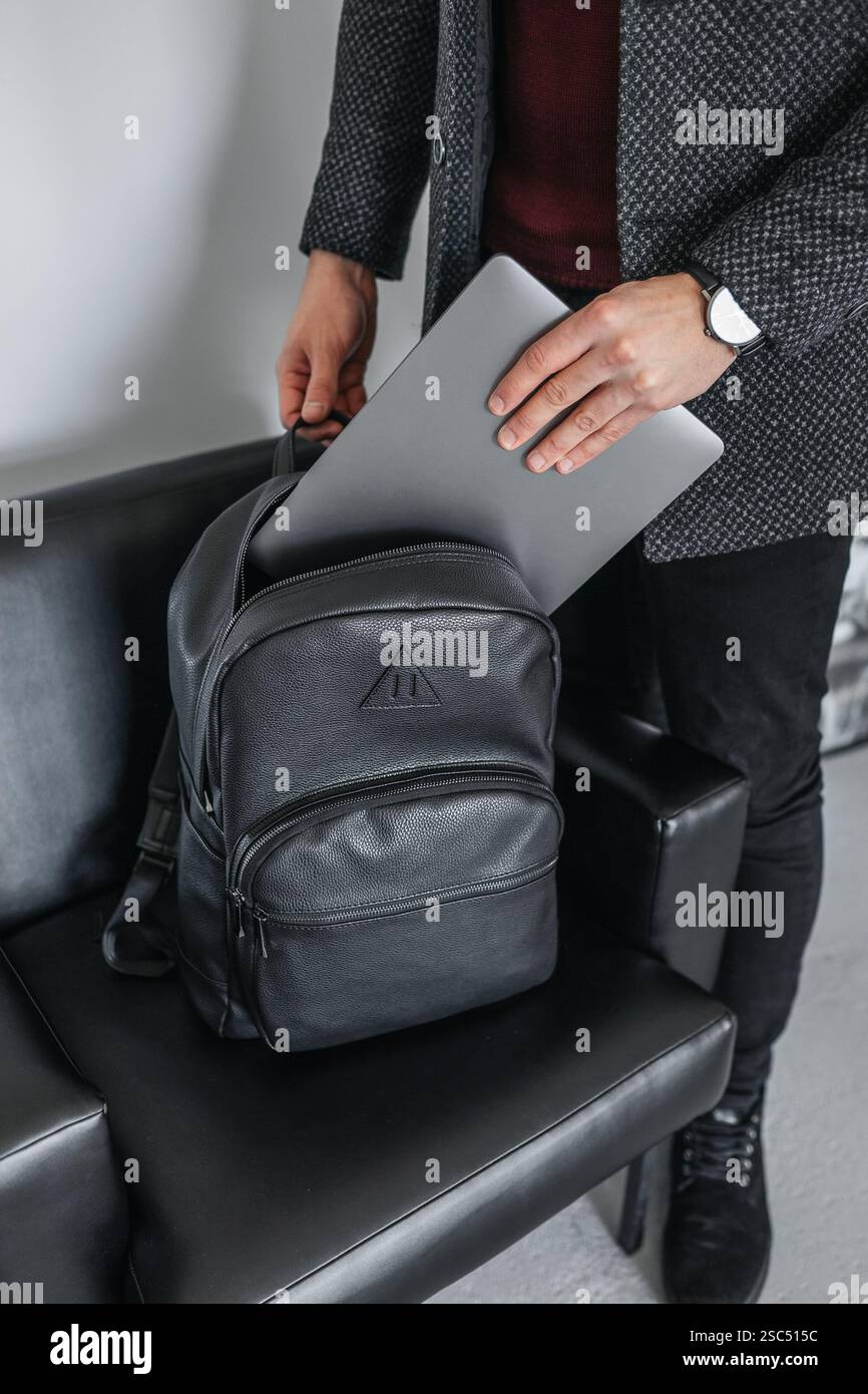 A man in a jacket puts his hand on a gray laptop in a stylish black leather backpack. Stock Photo
