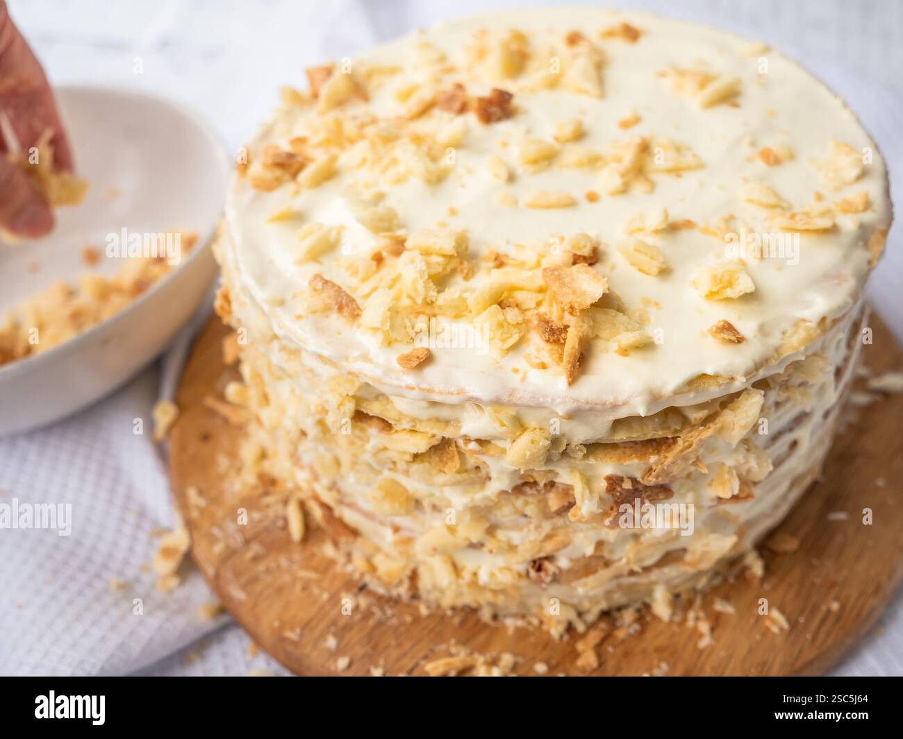 A hand sprinkling decorating crumbs over a pile of thin round layers spread with vanilla custard, a bowl of dough crumbs on a white waffle towel Stock Photo