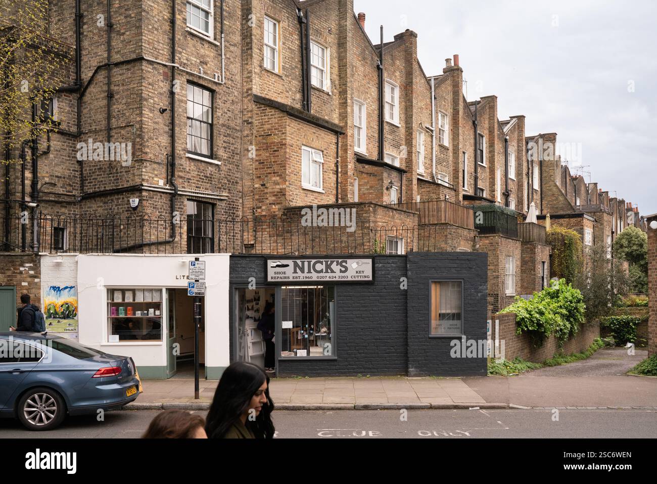 London, UK.  Shops (Nick's Shoe Repairs, LITE) on a street lined with terraced houses.  A car parks nearby. Stock Photo