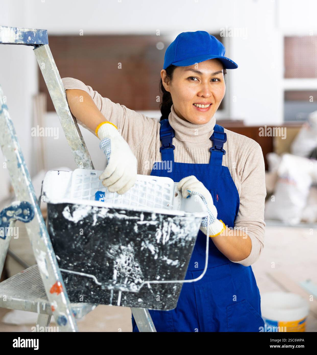 Woman painter in overalls with paint roller in building under ...