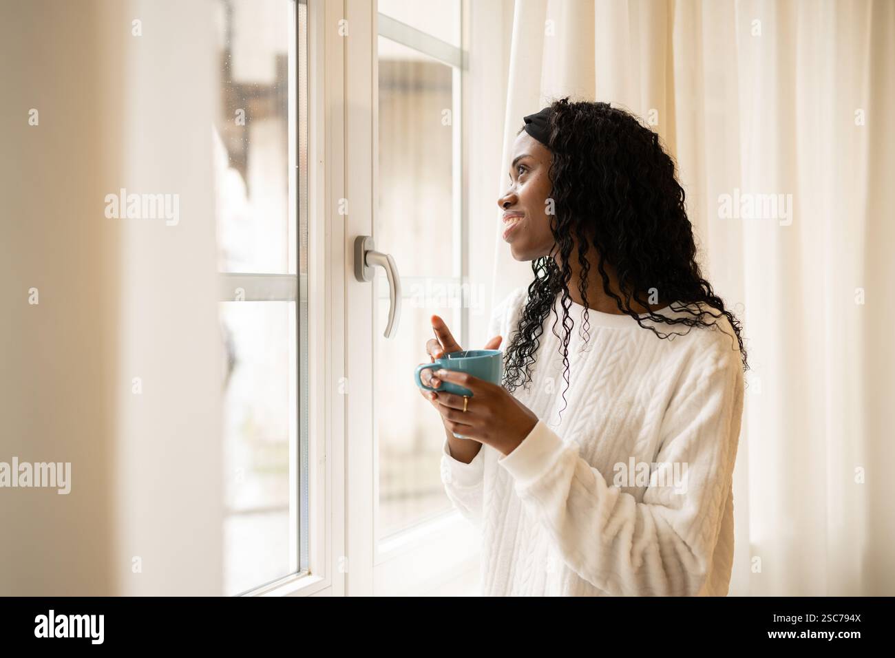 Young woman with long, curly hair holds a blue mug and gazes thoughtfully out a bright window, enjoying a quiet moment of reflection with her morning Stock Photo
