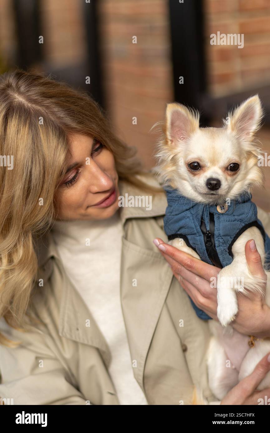 A woman dressed in a light jacket sits outdoors, gently holding a small dog wearing a denim coat. She smiles affectionately, enjoying the moment in a Stock Photo