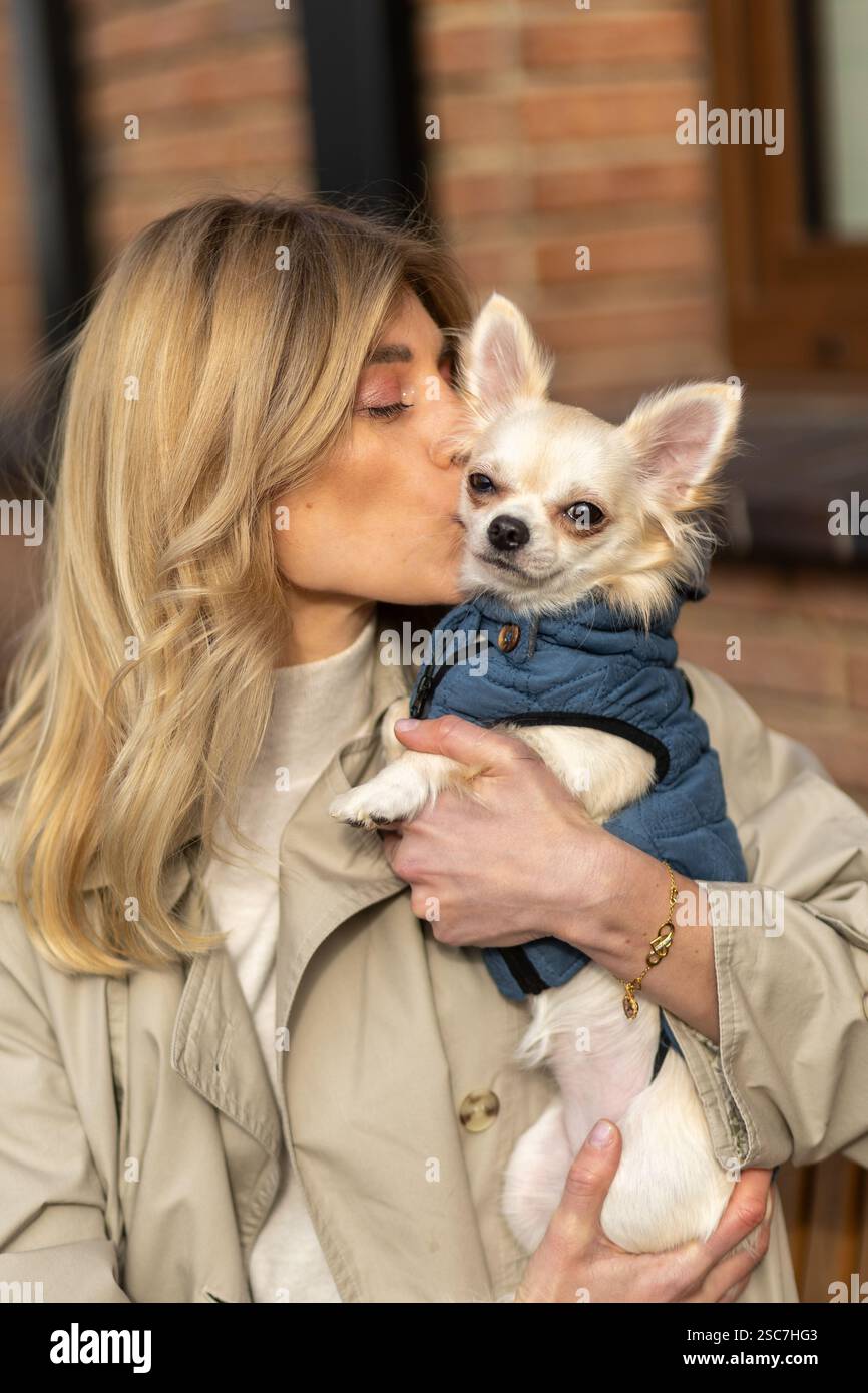 A woman wearing a light coat gently kisses her small dog dressed in a blue jacket. They are outside, enjoying a sunny day in a relaxed environment, sh Stock Photo