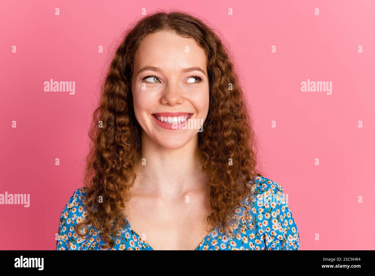 Portrait of a cheerful young woman with curly hair expressing surprise in a vibrant floral dress against a pink background Stock Photo