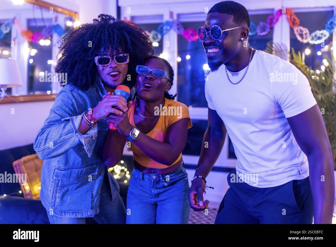 Excited friends holding microphone singing karaoke at home during night party Stock Photo