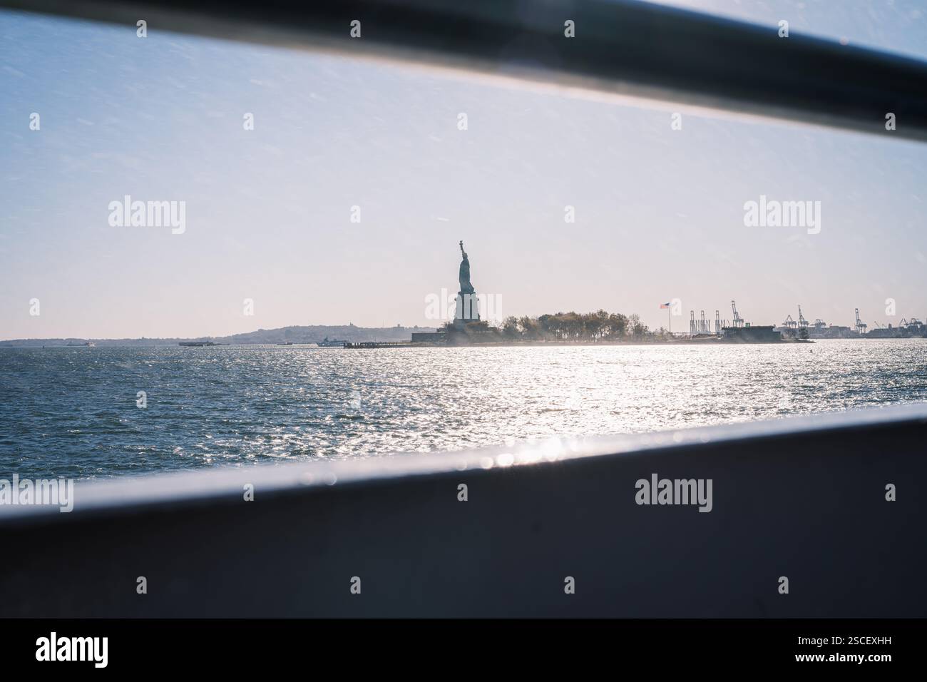 A stunning view of the Statue of Liberty from the ferry, shimmering waters, and an American flag against an industrial backdrop. Stock Photo