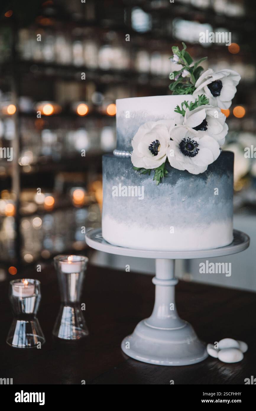 A beautifully crafted two-tier cake in shades of blue is decorated with delicate white flowers. Soft candlelight creates a cozy atmosphere in the back Stock Photo