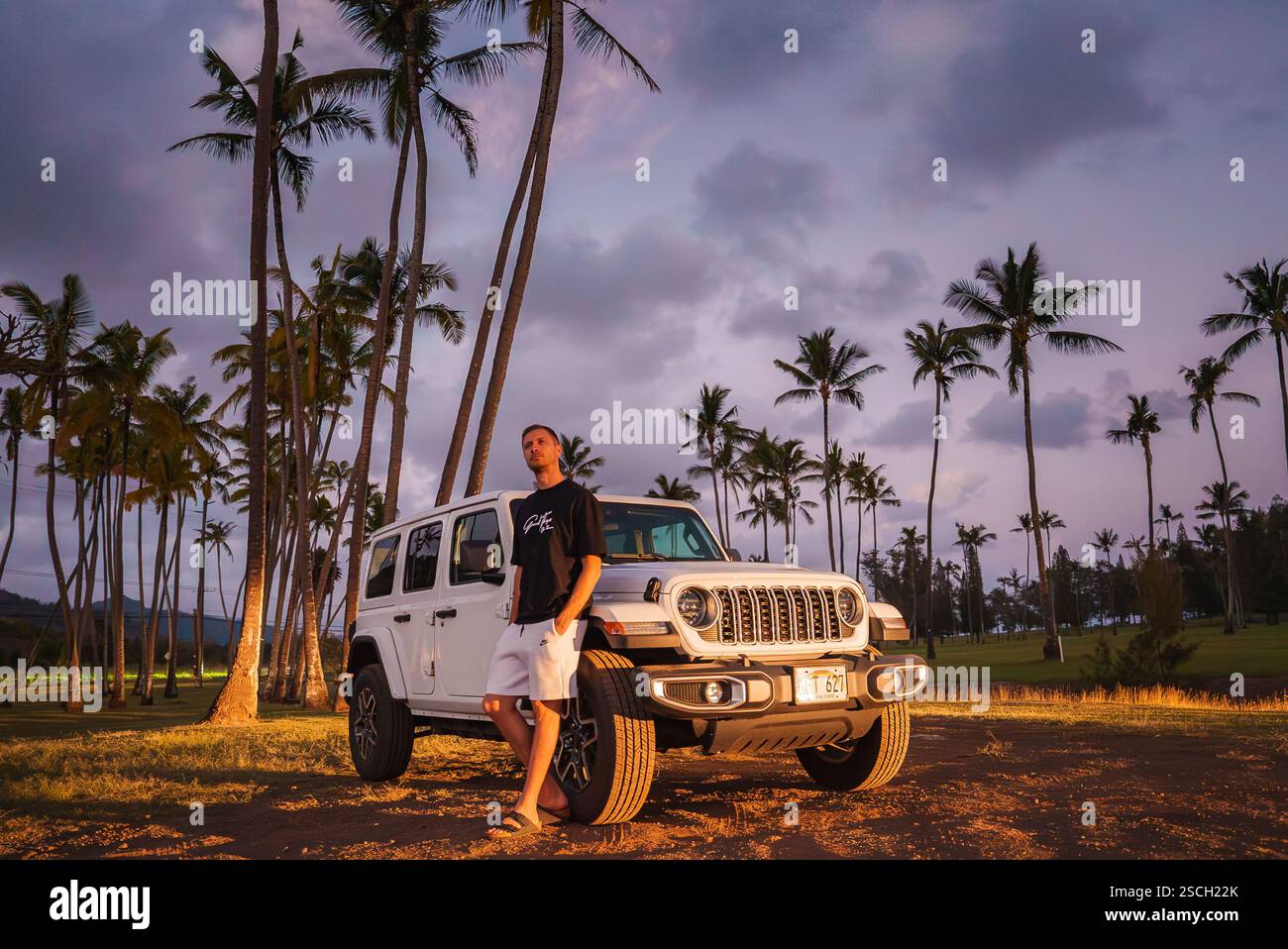 Man Standing by White Jeep Wrangler Among Palm Trees at Sunset Stock Photo
