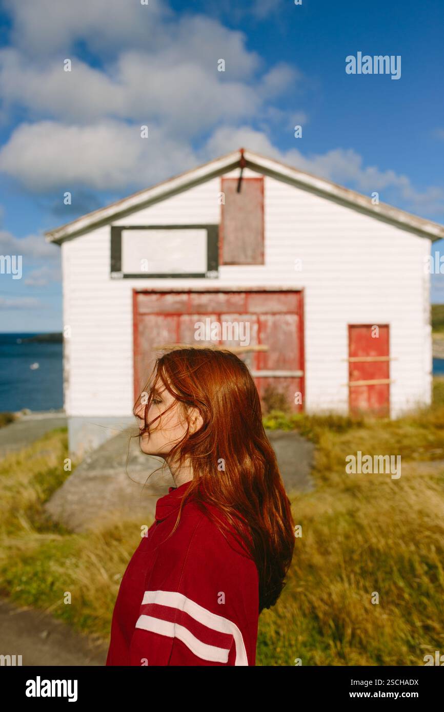 A young thoughtful woman with long red hair and eyes closed, wearing a red jacket, stands contemplatively in front of a white barn with red doors in F Stock Photo