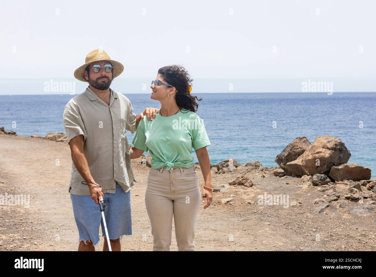 A blind man with a cane and a supportive woman walk side by side along a rocky coastal path. The serene ocean view in the background highlights their Stock Photo
