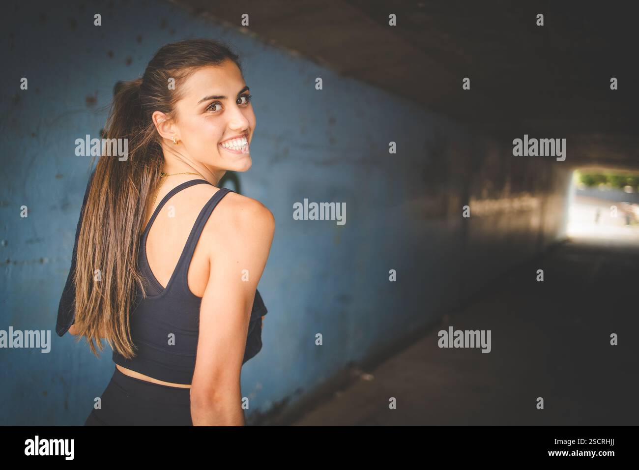 Young female athlete wearing black sportswear, engaging in various fitness poses outdoors against a modern industrial backdrop. Stock Photo