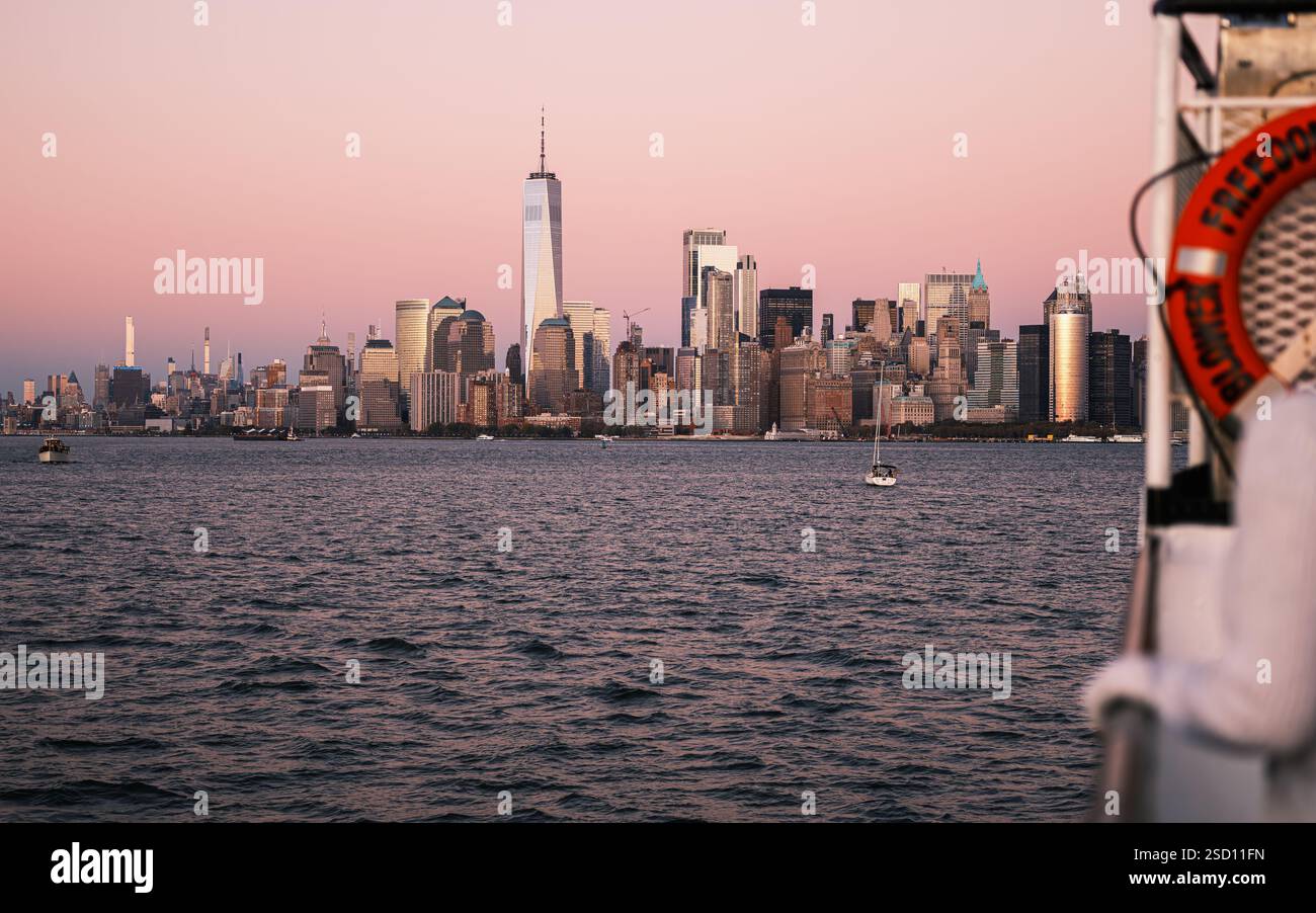 A golden hour view of Manhattan’s skyline from the Hudson River. One World Trade Center shines as boats drift on the water. Stock Photo
