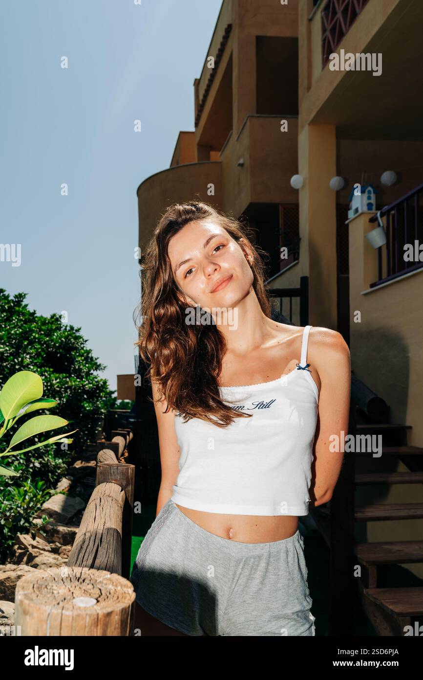 A cheerful young woman poses confidently outdoors by a wooden staircase, surrounded by lush greenery under the bright sun Stock Photo