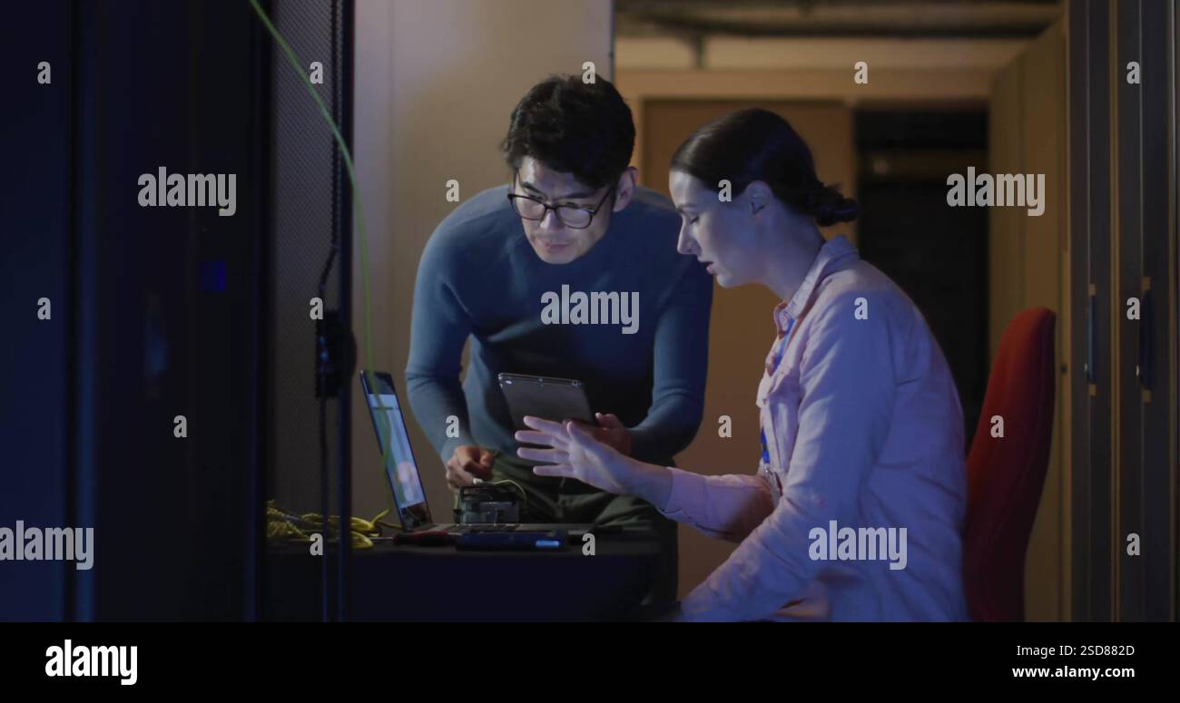 IT professionals collaborating on tablet and laptop in server room, focused on task Stock Photo