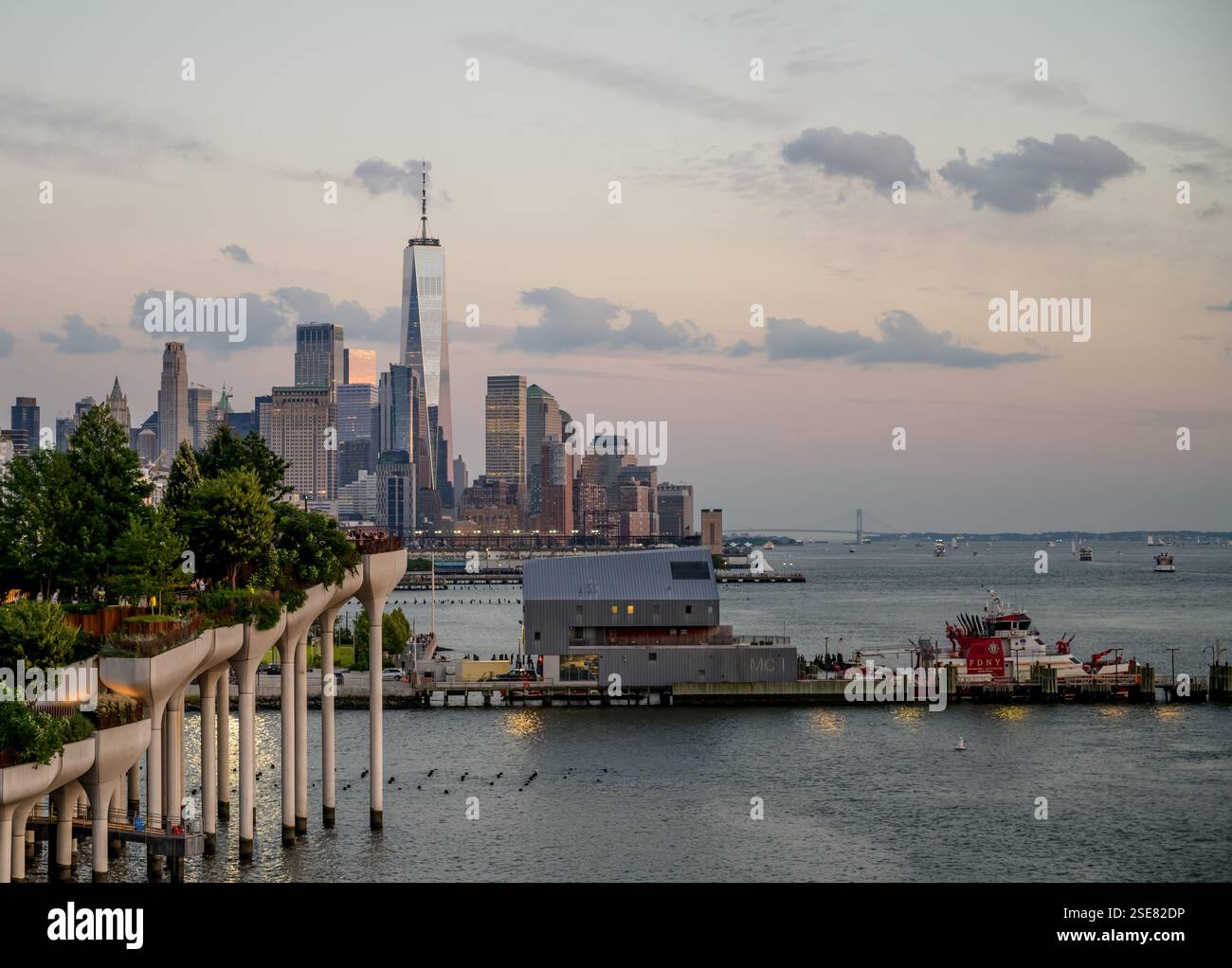 A beautiful view of the New York City skyline during dusk, with towering buildings silhouetted against the setting sun. People stroll along the waterf Stock Photo