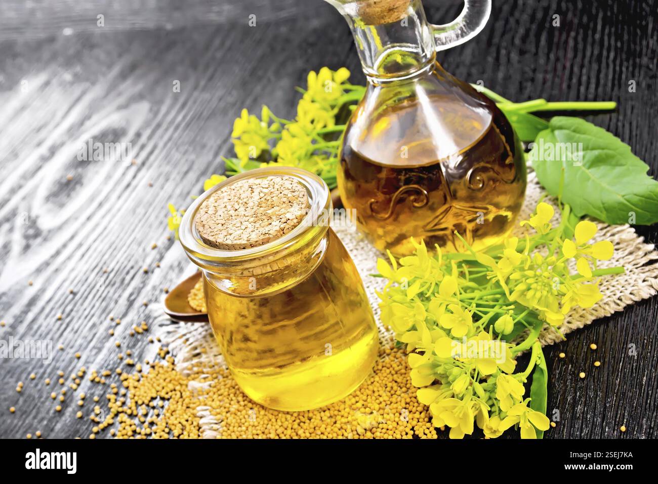 Mustard oil in a glass jar and decanter, mustard seeds on burlap, flowers and leaves on black wooden board background Stock Photo