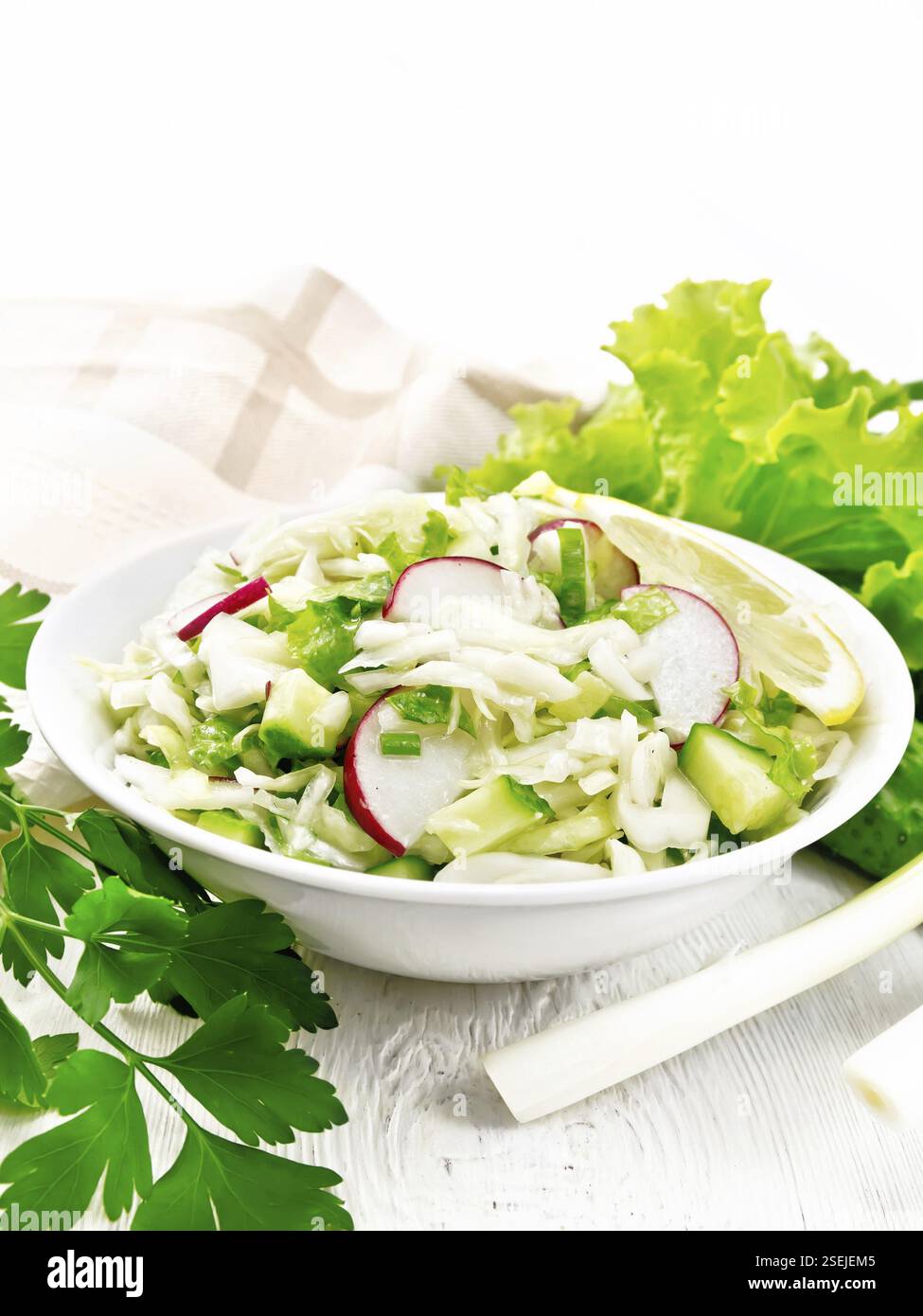 Salad of fresh cabbage, radish, lettuce, cucumber and green onion salad dressed with vegetable oil in a plate, napkin and parsley on wooden board back Stock Photo