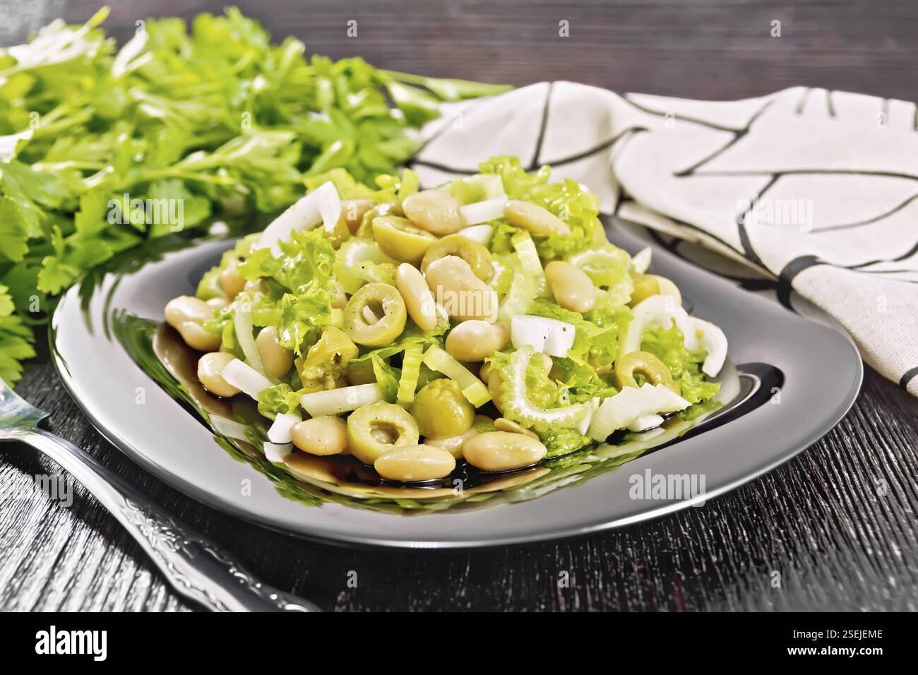 Salad of beans, olives, stalk celery, onion and lettuce, dressed with vegetable oil in a plate, napkin and fork on dark wooden board background Stock Photo