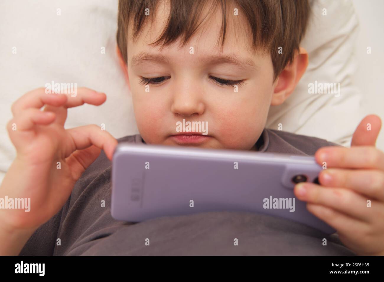 A close-up of a child using a mobile phone while lying in bed. A toddler exploring a mobile phone while resting. Kid aged three (three-year-old ) Stock Photo