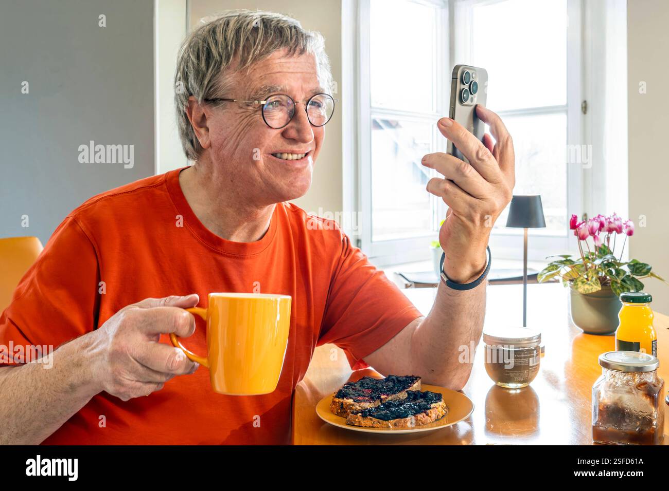 Rentner beim Frühstück zuhause, surft neben seinem Kaffee im Internet, Spätaufsteher, München, Februar 2025 Deutschland, München, Februar 2025, Seniorenfrühstück, Senior, Ü60, Mann liest beim Frühstück in seinem Handy, sitzt sonntags gemütlich im T-Shirt am Frühstückstisch, schaut in sein Apple Smartphone, Babyboomer, surft im Internet, am Wochenende Spätaufsteher, Langschläfer, Kaffeetrinker, Leben im Alter, allein leben, Single, Wohnzimmer, Innenaufnahme, Menschen, privat, Bayern, bayerisch, *** Pensioner at breakfast at home, surfs the Internet next to his coffee, late riser, Munich, Februa Stock Photo
