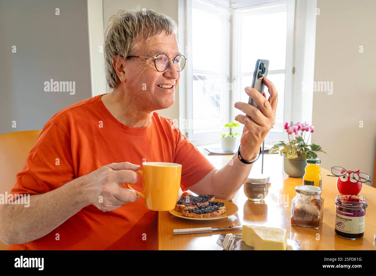 Rentner beim Frühstück zuhause, surft neben seinem Kaffee im Internet, Spätaufsteher, München, Februar 2025 Deutschland, München, Februar 2025, Seniorenfrühstück, Senior, Ü60, Mann liest beim Frühstück in seinem Handy, sitzt sonntags gemütlich im T-Shirt am Frühstückstisch, schaut in sein Apple Smartphone, Babyboomer, surft im Internet, am Wochenende Spätaufsteher, Langschläfer, Kaffeetrinker, Leben im Alter, allein leben, Single, Wohnzimmer, Innenaufnahme, Menschen, privat, Bayern, bayerisch, *** Pensioner at breakfast at home, surfs the Internet next to his coffee, late riser, Munich, Februa Stock Photo